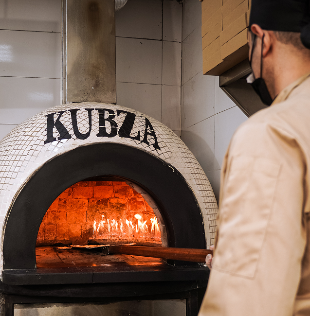 A baker wearing a uniform and face mask tending to a flaming stone oven at Kubza Bakery, preparing freshly baked bread.