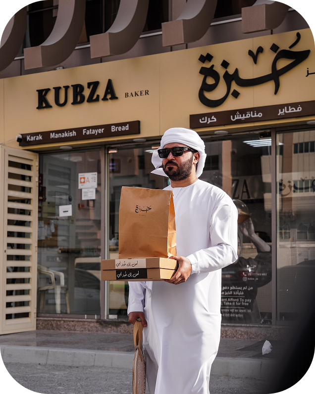 An Emirati man wearing traditional white attire and sunglasses walking outside Kubza Bakery, holding takeaway boxes and a paper bag with the bakery’s logo
